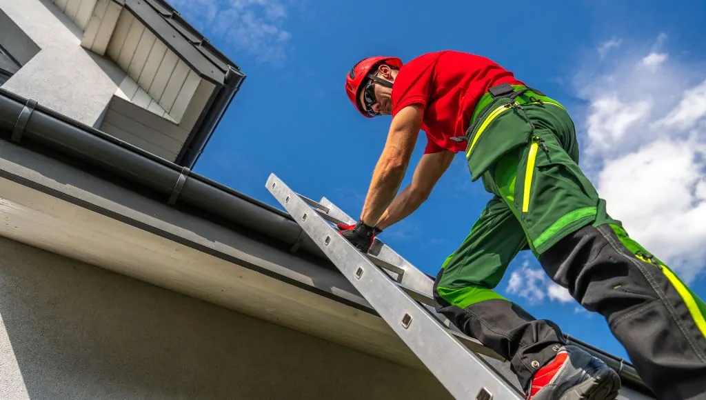 A professional roofer in a red shirt, green and black work pants, and a red hard hat, climbs a ladder to inspect a roof.