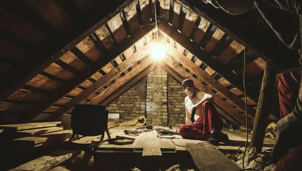 An adult wearing an N90 facemask, red pants, and a white top, fixes a damaged attic.