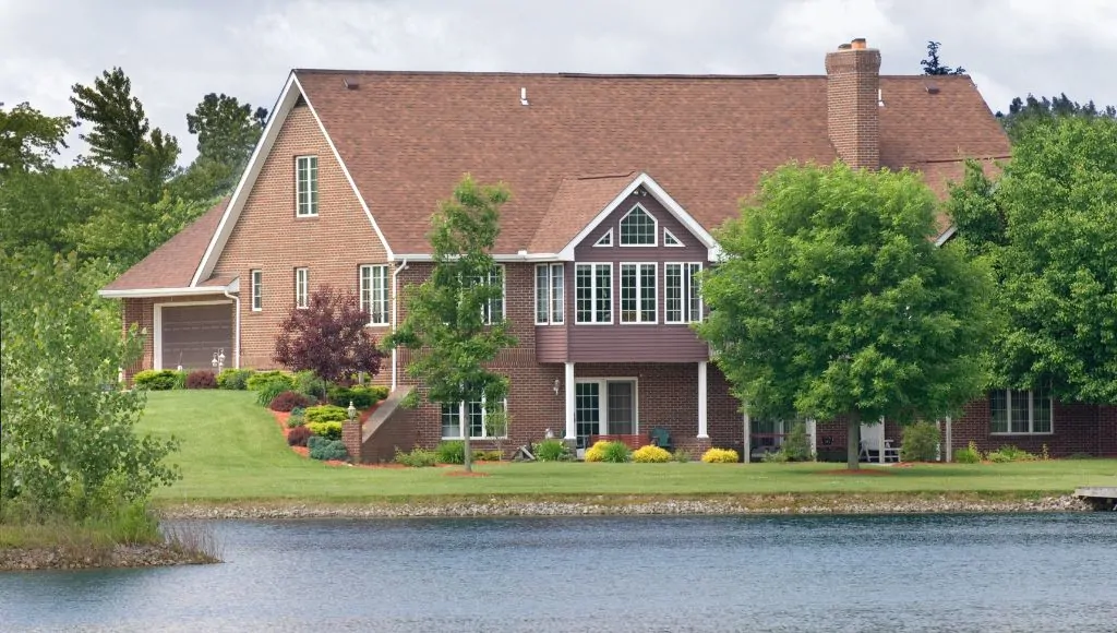 A brown brick lakeside home with manicured lawn and garden under a cloudy sky