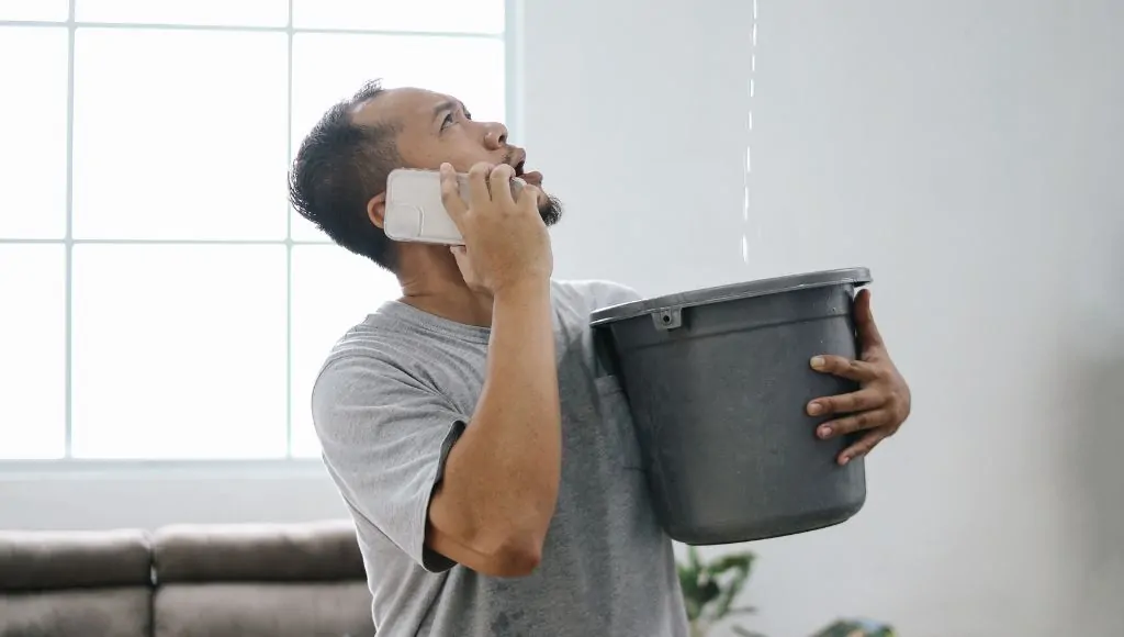 A man holds a bucket under a leak in his ceiling while on the phone calling in the professionals for help.