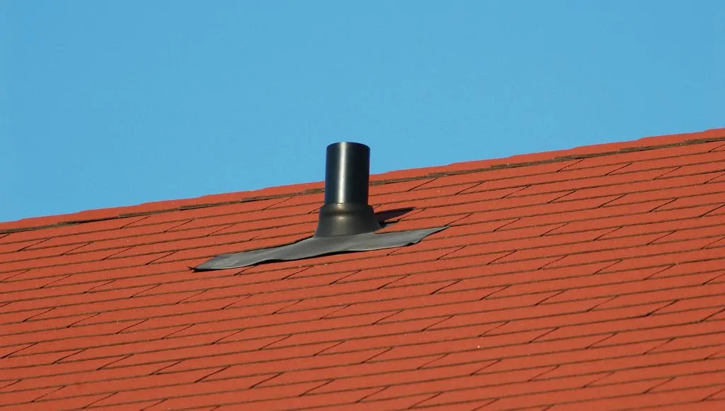 A rooftop covered in orange-red asphalt shingles, with a black vent pipe in the middle. The pipe seal seems to be lifting slightly.