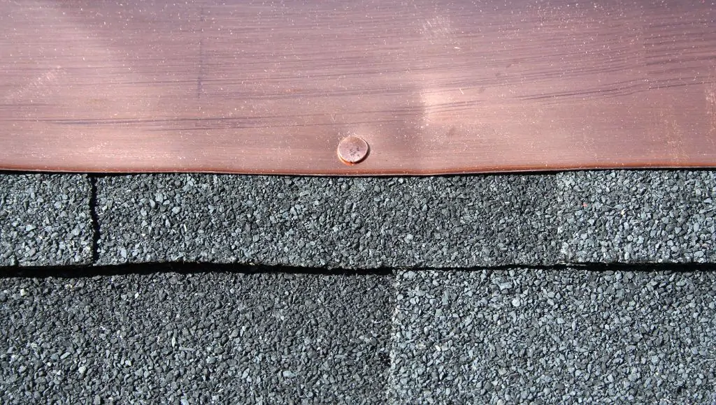 Close-up of copper colored flashing over dark grey shingles. The flashing has an obvious dent in it from damage.