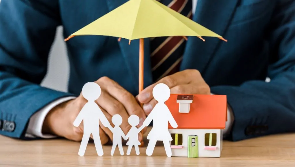 A conceptual image of a man in a dark blue suit holding a yellow umbrella to protect a small, colorful house and a paper family from the rain.